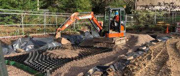 Mini excavator spreading gravel over cellular ground reinforcement grid on a construction site with forest backdrop